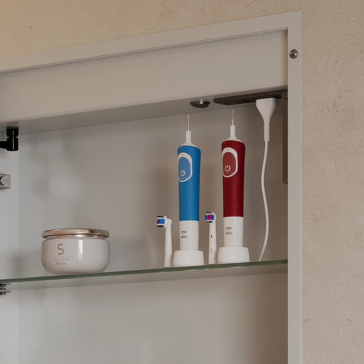 Two electric oral irrigators, one blue and one red, standing on white charging bases on a glass shelf inside a beige bathroom cabinet. An electric toothbrush and a small jar are also visible.
