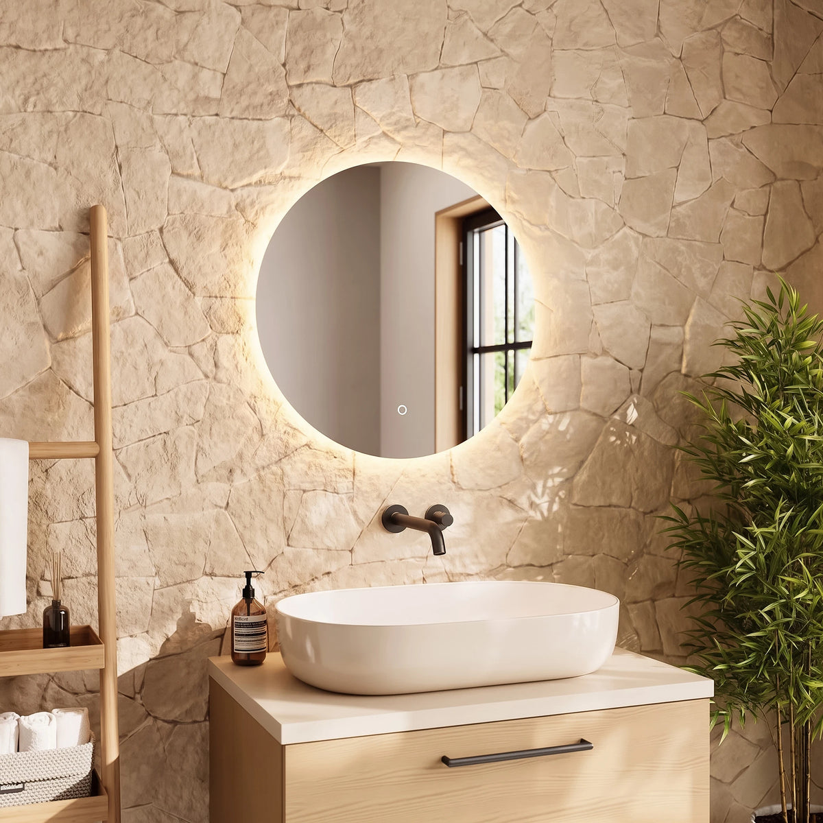 A modern bathroom scene featuring a round backlit LED mirror mounted on a textured stone wall above a light wood vanity with a white vessel sink. A wooden ladder shelf with toiletries stands on the left, and a potted bamboo plant is on the right. #Size_⌀600mm