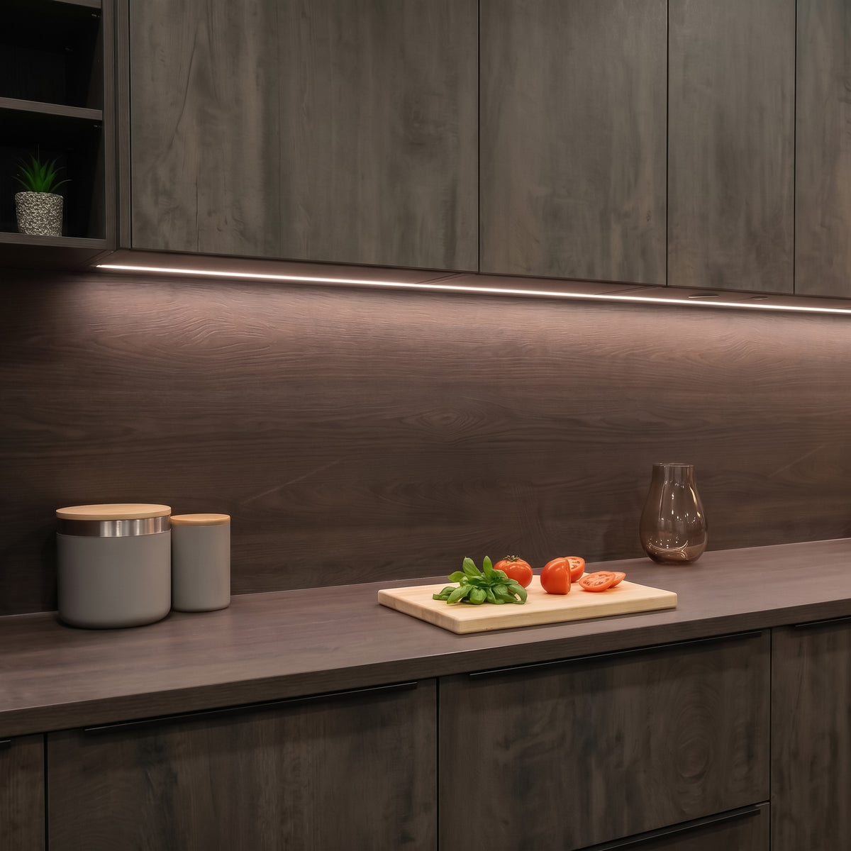A modern kitchen scene with dark wood cabinets, a matching countertop, and a dark wood backsplash illuminated by under-cabinet LED lighting. A small cutting board with tomatoes and basil sits on the counter, along with two gray canisters and a glass vase. #Finish_Black