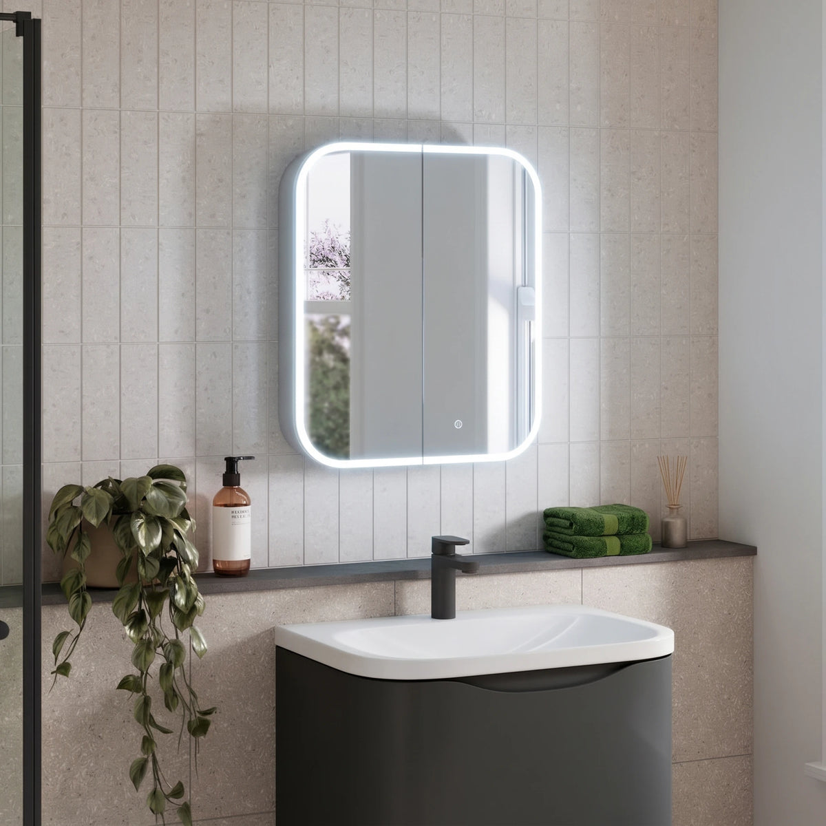A modern bathroom scene featuring a rectangular LED-backlit medicine mirror mounted on a light beige tiled wall above a dark gray and white vanity with a black faucet. A green trailing plant, a brown bottle, and a folded green towel are arranged on the shelf. #Size_600mm x 700mm
