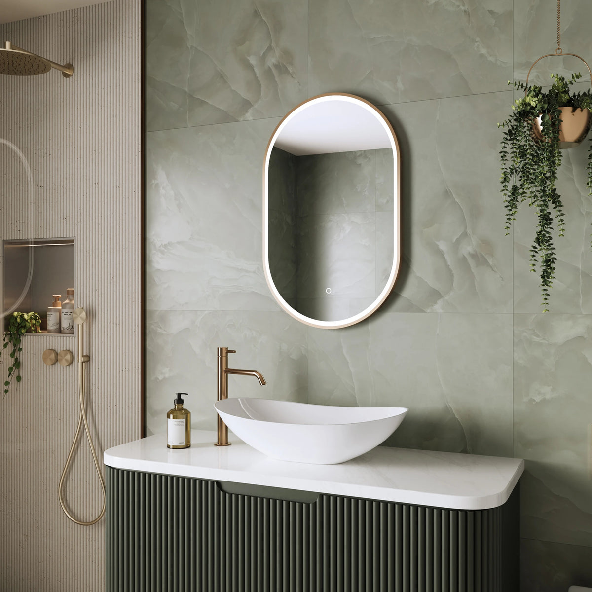 A modern bathroom vanity setup with a white oval vessel sink, a gold faucet, a ribbed dark green cabinet, and an oval backlit mirror mounted on a light green marble-look tiled wall. A gold shower head and hand shower are visible on the left, and a hanging green plant decorates the right side. #Finish_Brushed Brass