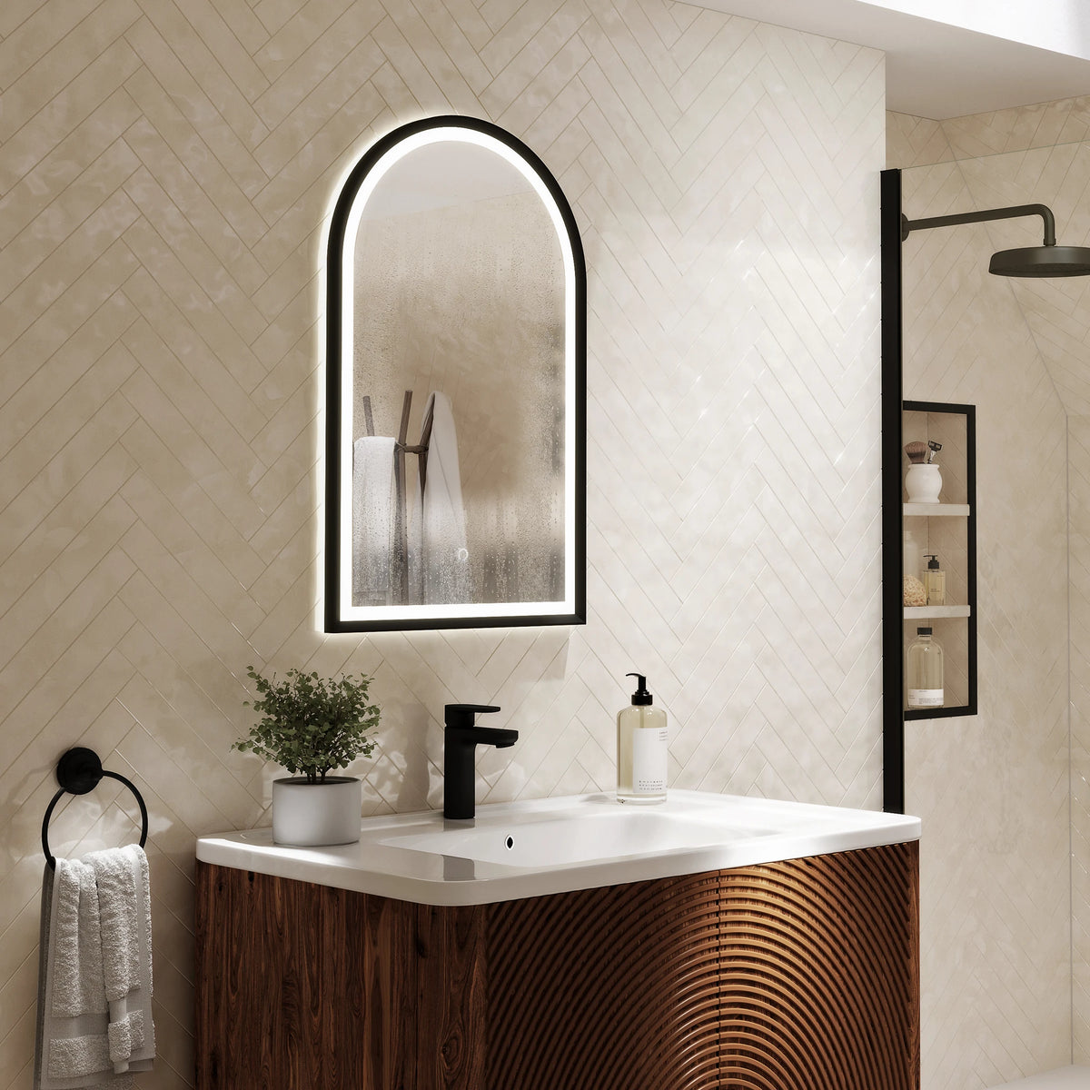An illuminated arched bathroom mirror with a black frame hangs above a white sink and a dark wood vanity in a modern beige tiled bathroom. #Finish_Black