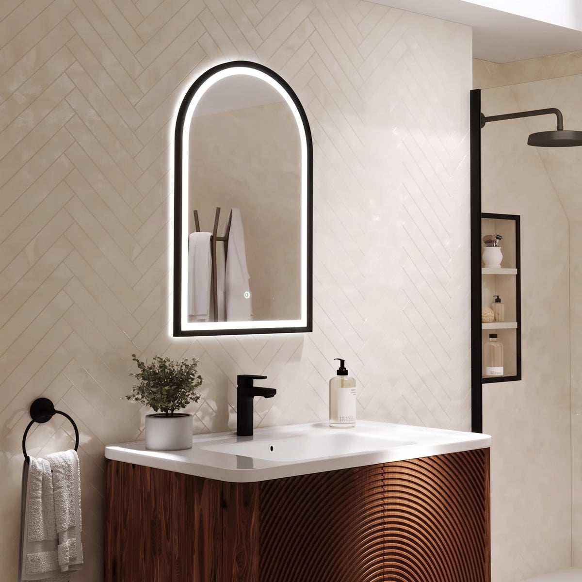 A modern bathroom vanity setup with a white rectangular sink, dark wood cabinet, black faucet, and a large arched backlit mirror mounted on a beige tiled wall. A small plant, soap dispenser, towel ring, and shower area are visible in the room. #Finish_Black