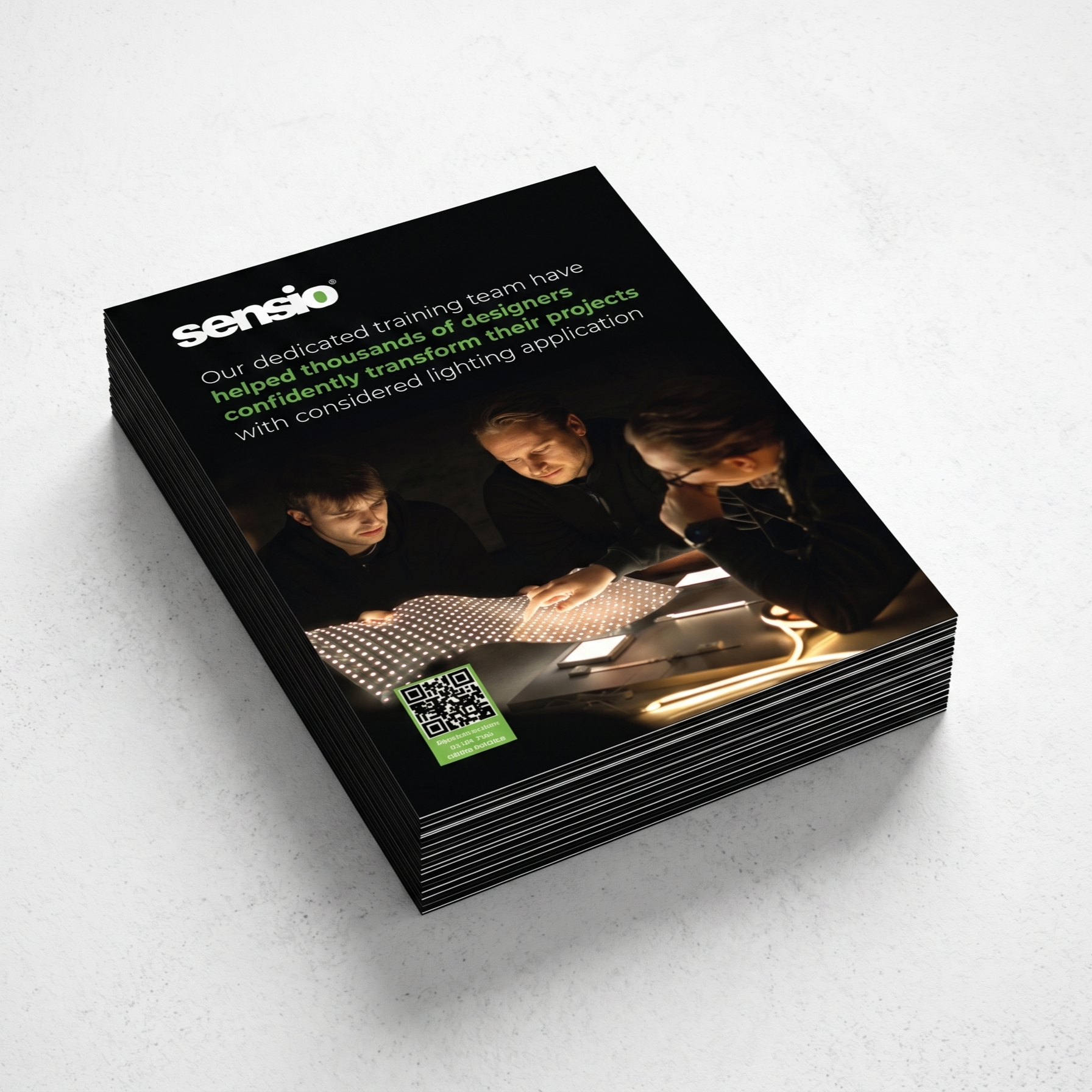 A stack of printed Sensio brochures or training program folders photographed on a light gray surface. The top brochure is angled to show the front cover, which is predominantly black with white and green text, a photo of three people working at a light fixture, and a green QR code.